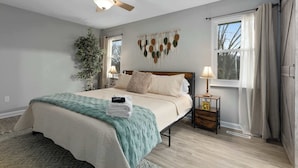 Une chambre avec un lit, un ventilateur de plafond, une décoration murale en macramé, une table de chevet avec une lampe et une fenêtre avec des rideaux.