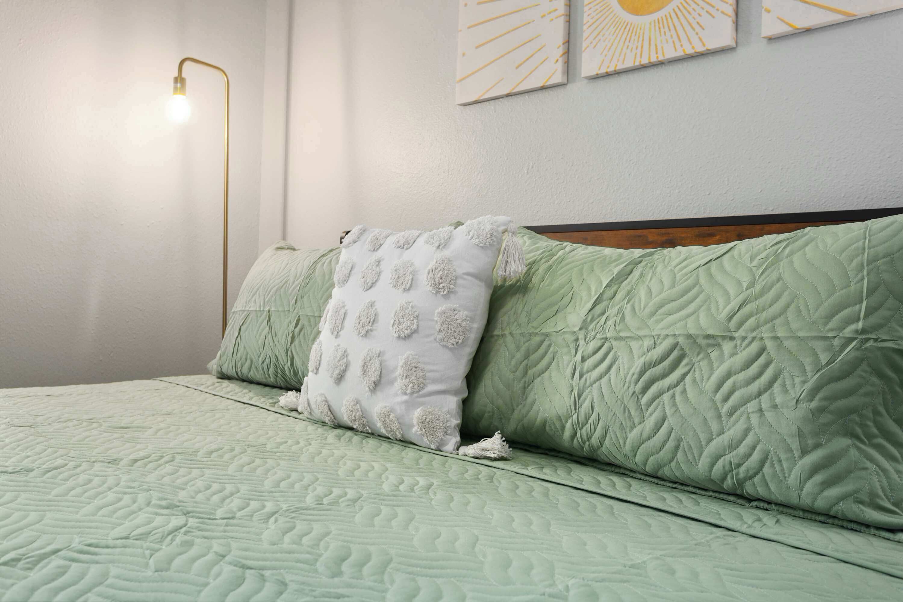 A bed with a green quilted bedspread, a white pillow with tassels, and a gold floor lamp.