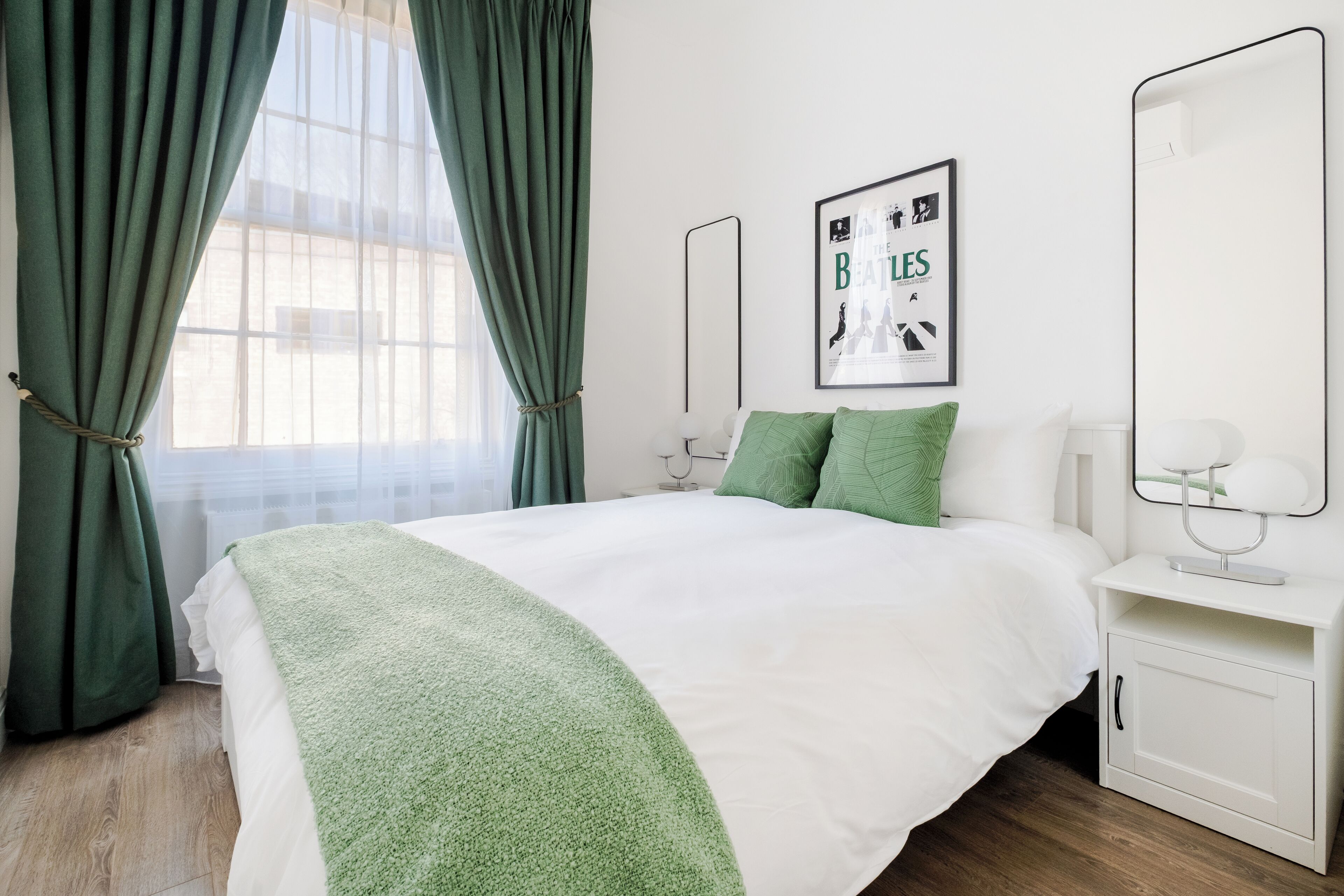 A neatly arranged bedroom with a large bed, green curtains, a framed poster on the wall, and a bedside table with a lamp.