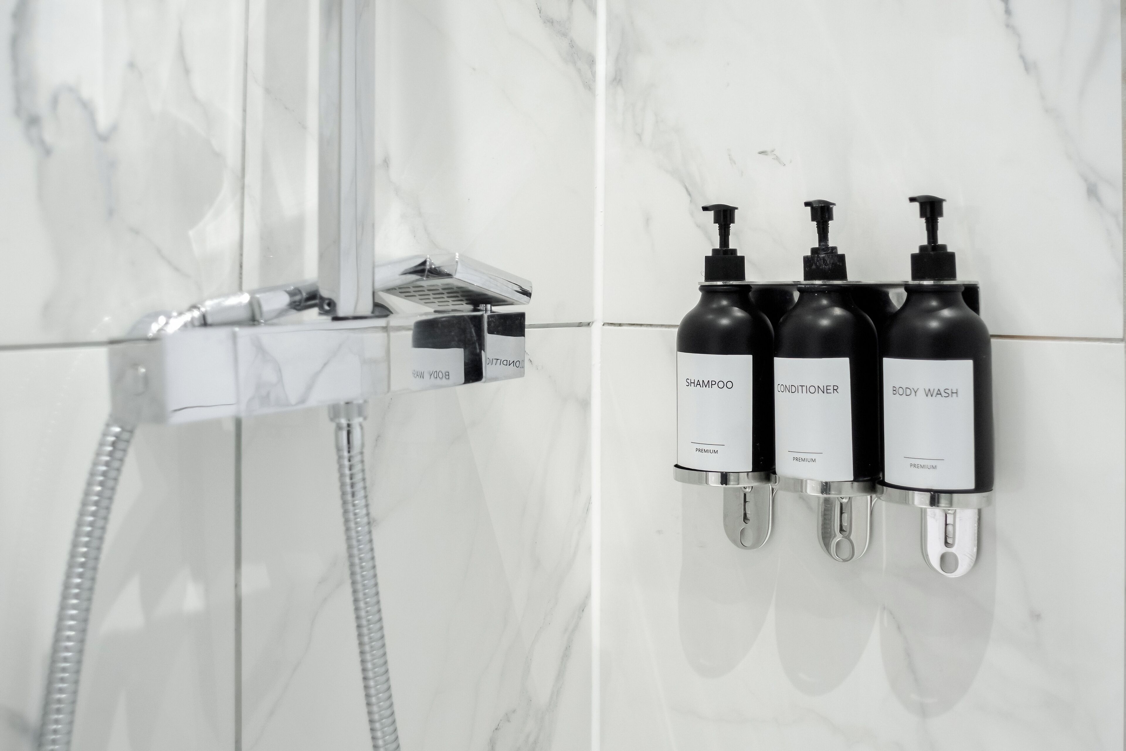 A bathroom with a shower, a hand dryer, and three bottles of toiletries labeled shampoo, conditioner, and body wash.