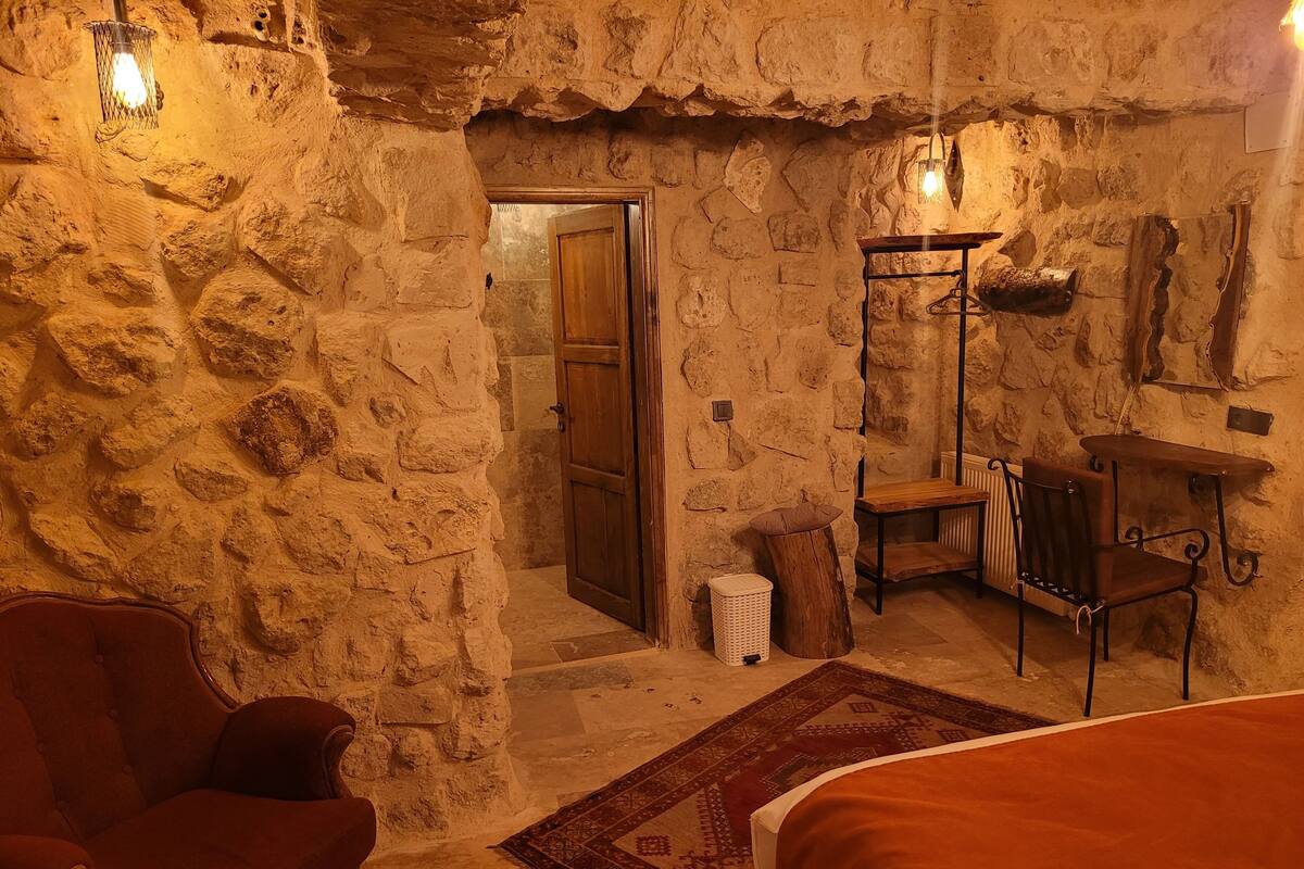 A rustic stone-walled room with a wooden door, a red armchair, a small table with a chair, and a hanging light fixture.
