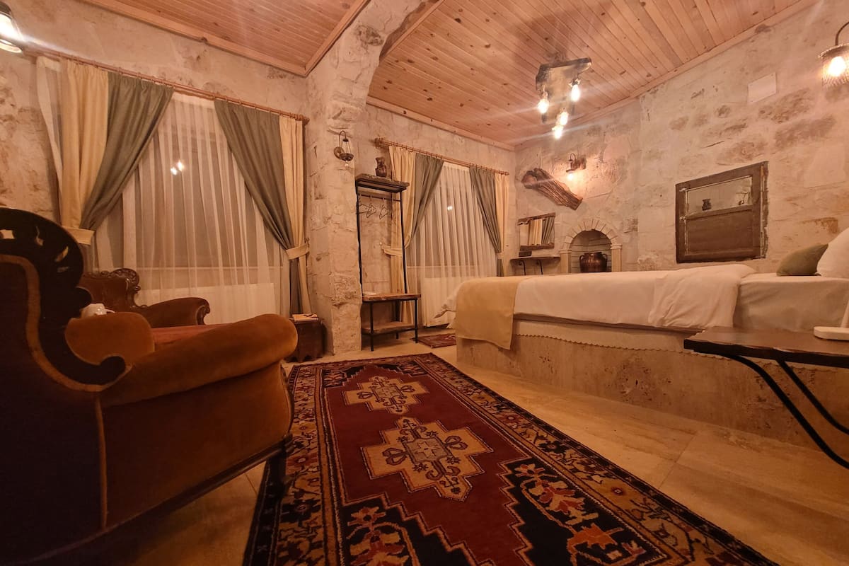 A cozy room with a wooden ceiling, a bed with a white bedspread, a brown armchair, a patterned rug, and a small table.