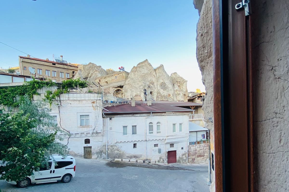 A street view with a white car parked on the left, buildings with traditional architecture, and a rocky hill in the background.