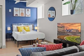 A modern hotel room with a bed, a TV, a sofa, and a painting of a racetrack.
