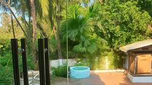 A glass door leading to a patio area with a pool, surrounded by lush greenery and palm trees. - The Grantley Hotel (Alibag)