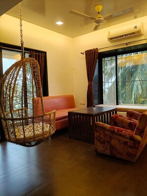 A room with a hanging chair, a sofa, a table, and a ceiling fan. - The Grantley Hotel (Alibag)