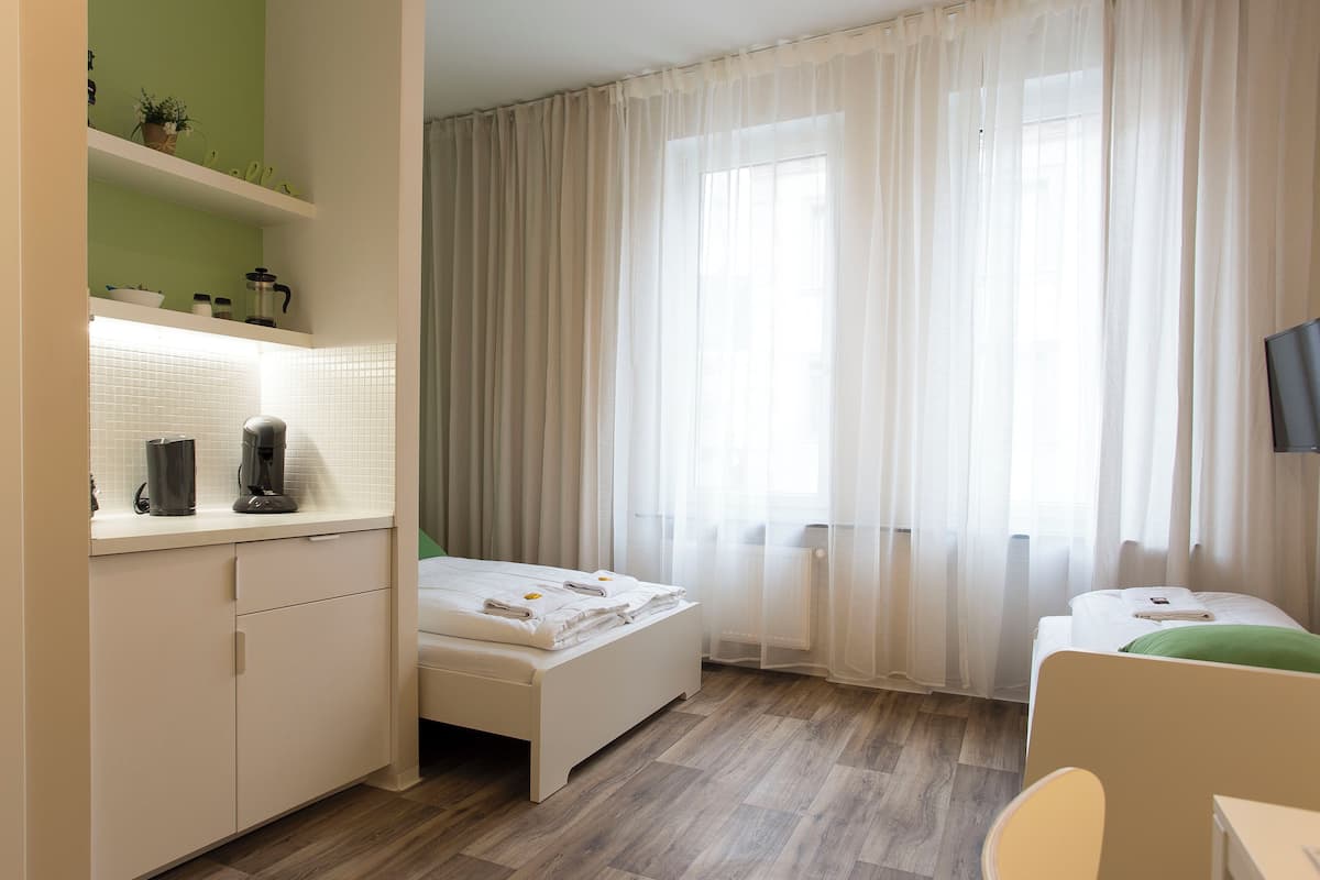A hotel room with a bed, a small desk with a chair, a TV, a window with sheer curtains, and a kitchenette area with a coffee maker and a kettle.