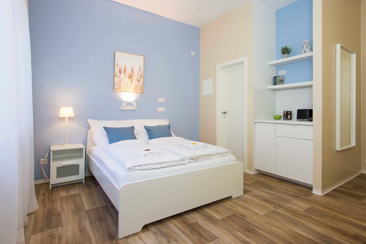 A bedroom with a bed, bedside table, lamp, and a small kitchenette area.