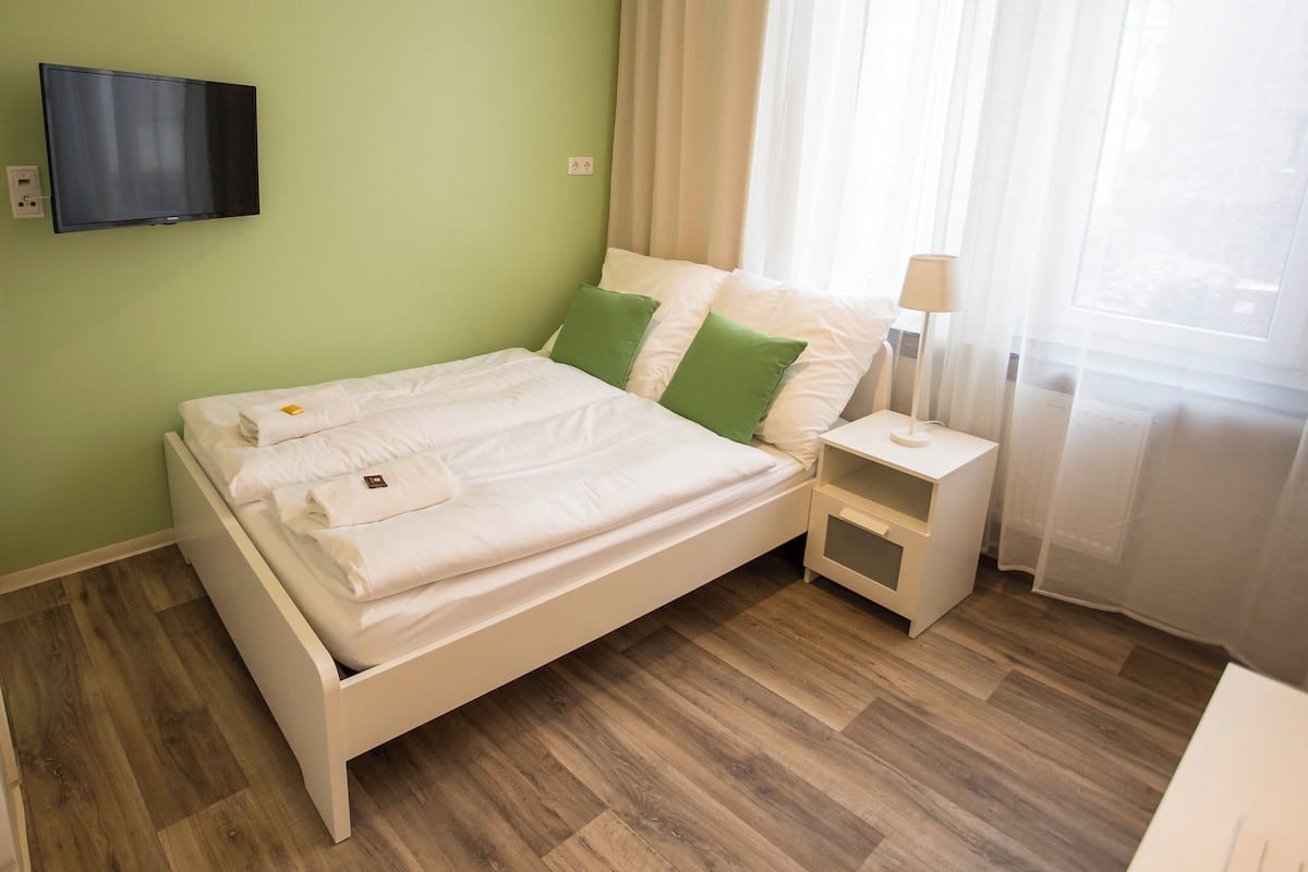 A neatly made bed with white linens and green pillows, a bedside table with a lamp, and a flat-screen TV mounted on the wall.
