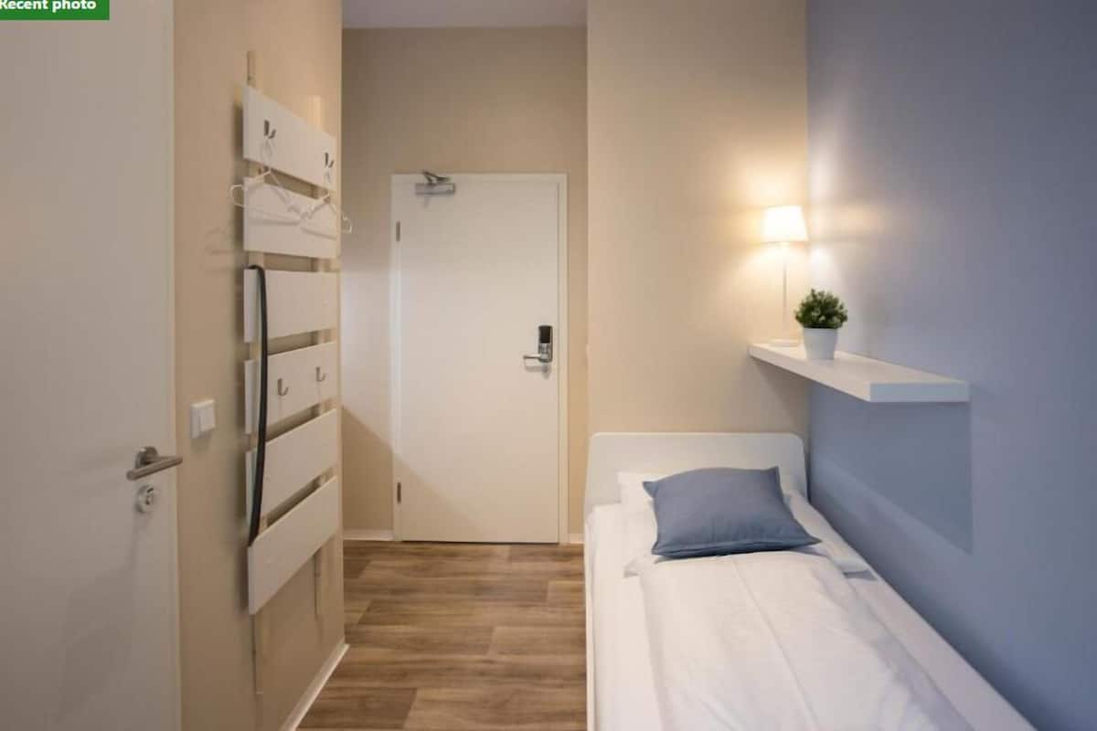 A bedroom with a bed, a bedside table with a lamp, a wall-mounted shelf, and a door with a coat rack.