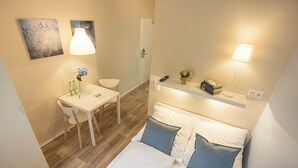 A hotel room with a bed, bedside table, chair, desk, and a small shelf with books and a plant. - GuestHouse Köhn51 (Nuremberg)