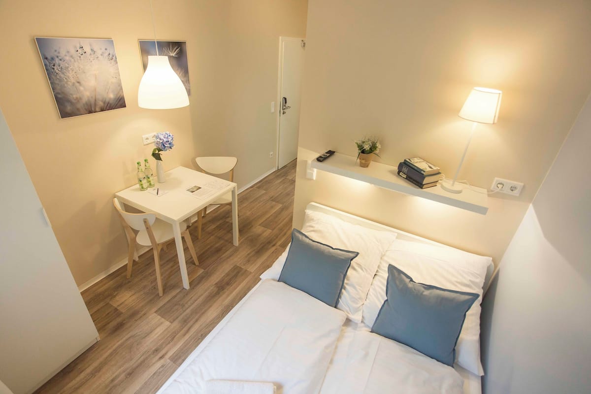 A hotel room with a bed, bedside table, chair, desk, and a small shelf with books and a plant.