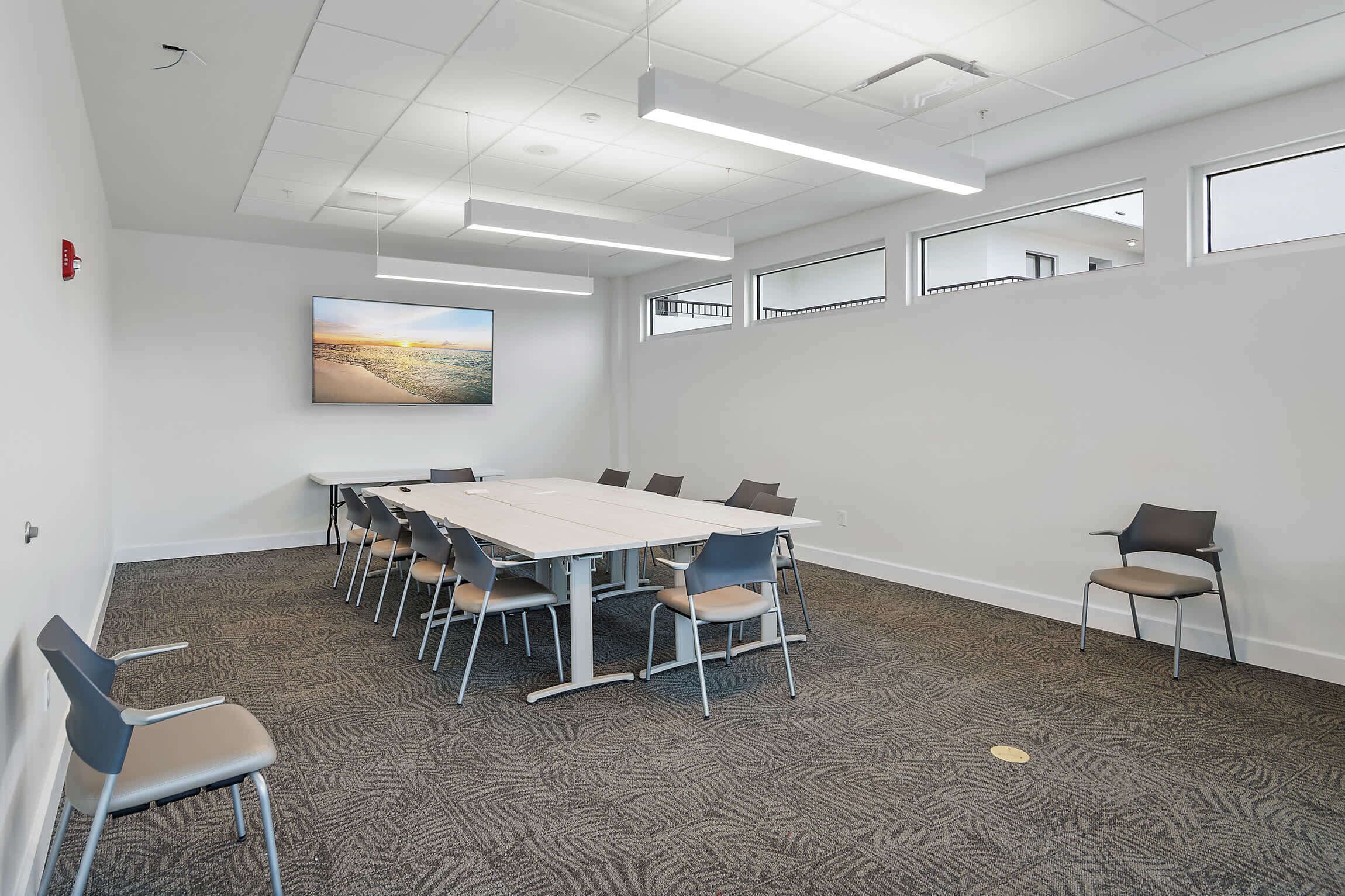 A conference room with a large table, chairs, carpeted floor, white walls, and a painting.