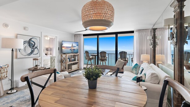 A spacious living area with a wooden dining table, white sofas, and a large window offering a view of the ocean. - Seagrove Beachfront Condo with Incredible Views (Santa Rosa Beach)