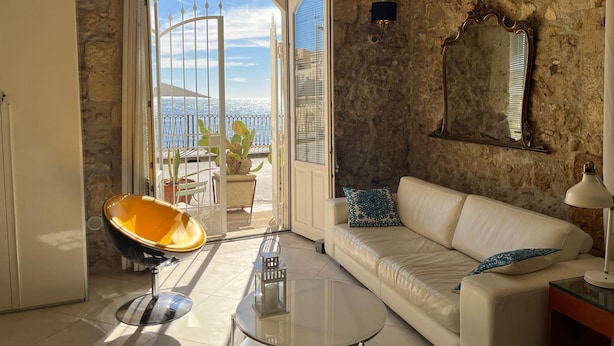 Una sala de estar con un sofá blanco, una mesa de centro de vidrio, una silla amarilla y vistas al mar a través de un ventanal.
