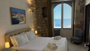 Habitación de hotel con cama, dos sillas, un cuadro y vista al mar a través de una ventana.