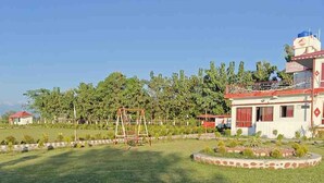 A grassy area with a swing set, a building with a red roof, and a small structure with a red roof in the background. - Amantran Hotel & Resort (Alipurduar)