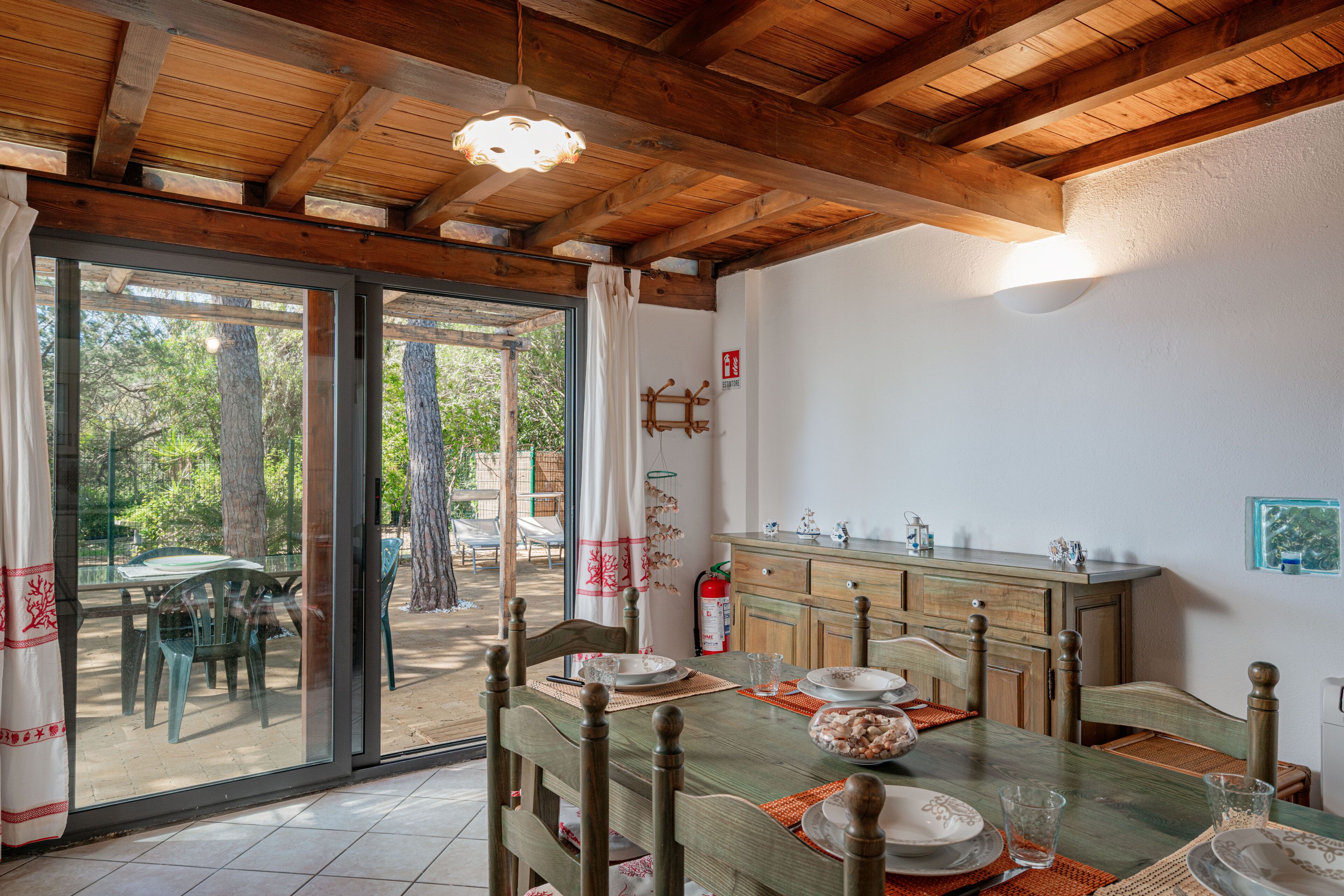 A dining area with a wooden table set for four, a fire extinguisher, and a view of the outdoors through glass doors.