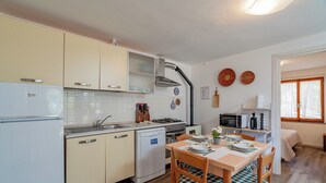 A compact kitchen with a dishwasher, stove, and microwave. There is a dining table with chairs and a window with curtains. - Casa Donatella (Orosei)