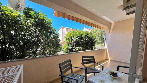 A balcony with a table and chairs, a white satellite dish, and a view of greenery and buildings. - Apartman Portos (Crikvenica)