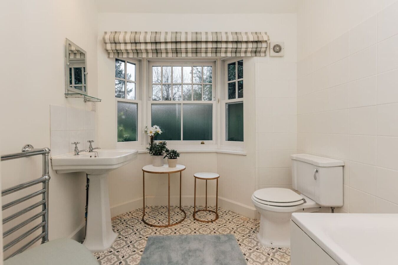 A bathroom with a white sink, toilet, and bathtub. There is a window with a striped valance, and two small round tables with plants.