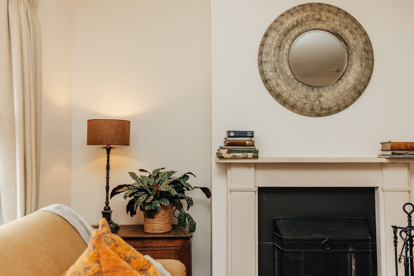 A cozy living room with a fireplace, a lamp, a plant, and a stack of books on the mantel.