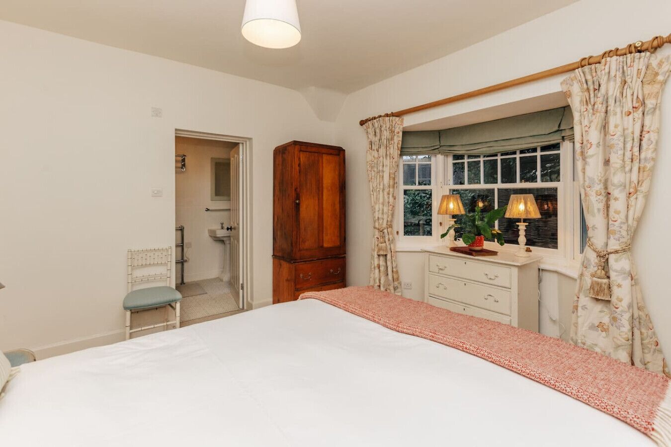 A bedroom with a bed, a wooden wardrobe, a vanity with a mirror, a window with curtains, and a small chair.