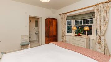 A bedroom with a bed, a wooden wardrobe, a vanity with a mirror, a window with curtains, and a small chair.