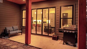 A covered patio with a sliding glass door leading to a kitchen area, a barbecue grill, and a seating area with a sofa and chairs.