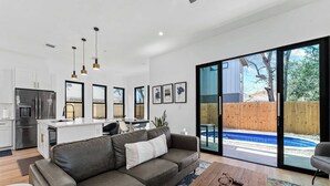 A modern kitchen with stainless steel appliances, a dining area with a white table and chairs, a gray sofa with a white pillow, and a wooden coffee table. - Downtown ATX - Unit A | Modern Home | Pool (Austin)