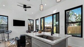 A modern kitchen with a large island, black bar stools, a ceiling fan, and large windows offering views of the backyard.