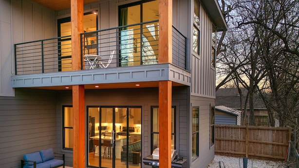 A modern two-story house with a balcony, a patio with outdoor seating, and a visible kitchen area.
