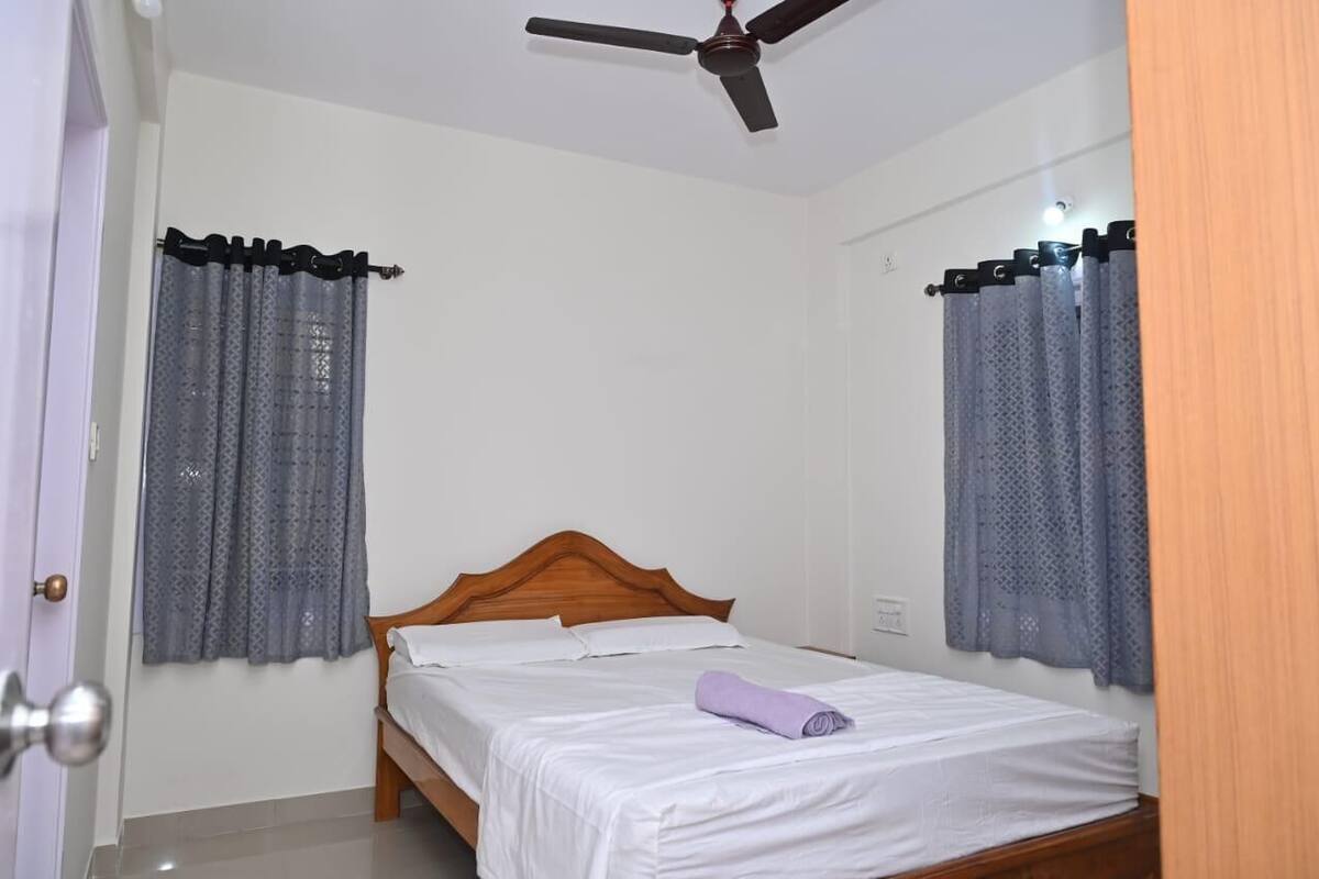 A bedroom with a wooden bed, white bedding, a ceiling fan, and lace curtains.