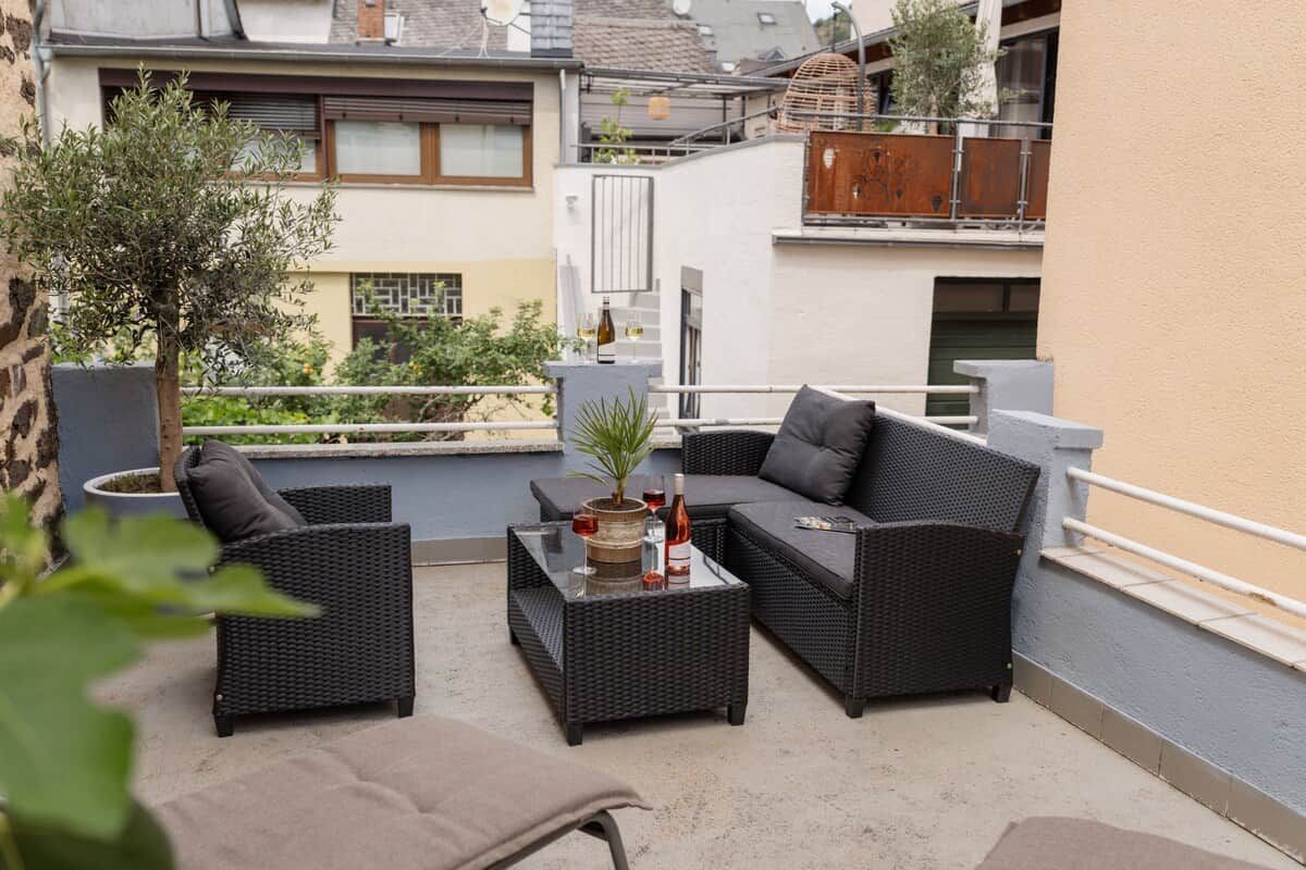 An outdoor seating area with wicker furniture and a glass coffee table, surrounded by plants and a view of neighboring buildings.