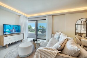A modern hotel room with a large TV, a sofa, a round ottoman, and a balcony with outdoor furniture.