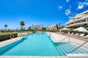 A swimming pool with clear blue water, surrounded by lounge chairs and umbrellas, with a building in the background.