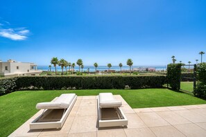 Una terraza con dos tumbonas, un césped cuidado y vistas al mar y palmeras.