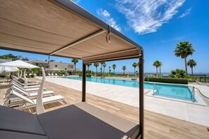 A poolside area with sun loungers and umbrellas, a clear blue sky, and palm trees.