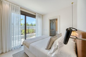 A bedroom with a large bed, a window with sheer curtains, a view of the outdoors, and a bedside table with a lamp.