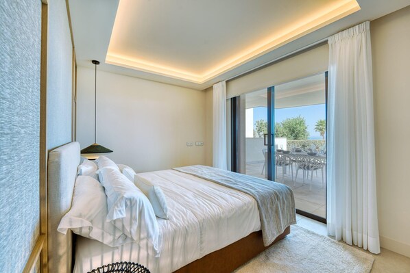 Une chambre à coucher comprenant un lit, une table de chevet, une lampe et donnant sur une terrasse extérieure avec une table et des chaises.