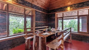 A dining area with a wooden table set for four, benches, and large windows with a view of greenery outside. - Alaya Stays Suro Woodbine Chalet Cottage (Kandaghat)