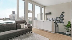 Un salón moderno con un sofá gris, ventanas amplias, un radiador blanco y un adorno de pared hexagonal.