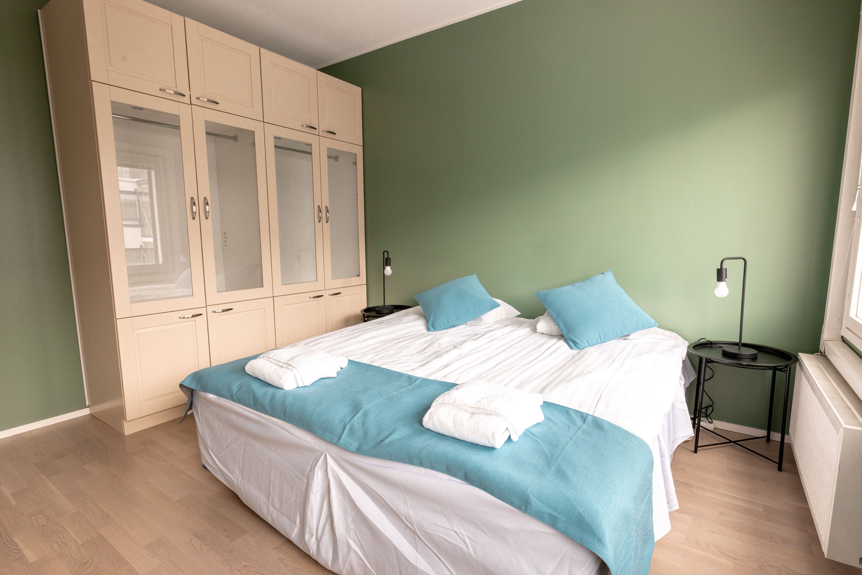 A neatly made bed with white and blue bedding, a large wardrobe with glass doors, and a bedside table with a lamp.