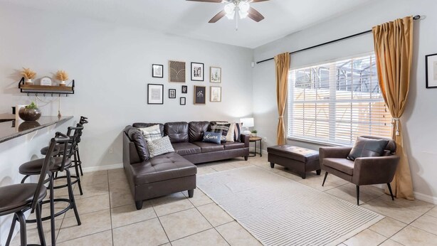 A modern living room with a dark brown leather sofa, a coffee table, and a dining area with bar stools. There is a ceiling fan, a large window with curtains, and framed artwork on the wall. - Kissimmee 4BR Retreat | Resort Access & Pool (Kissimmee)