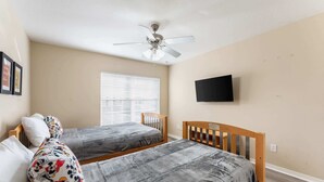 Une chambre avec deux lits, un ventilateur de plafond, un téléviseur fixé au mur et une fenêtre avec des stores.