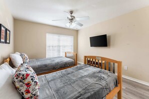 Une chambre avec deux lits, un ventilateur de plafond, un téléviseur fixé au mur et une fenêtre avec des stores.