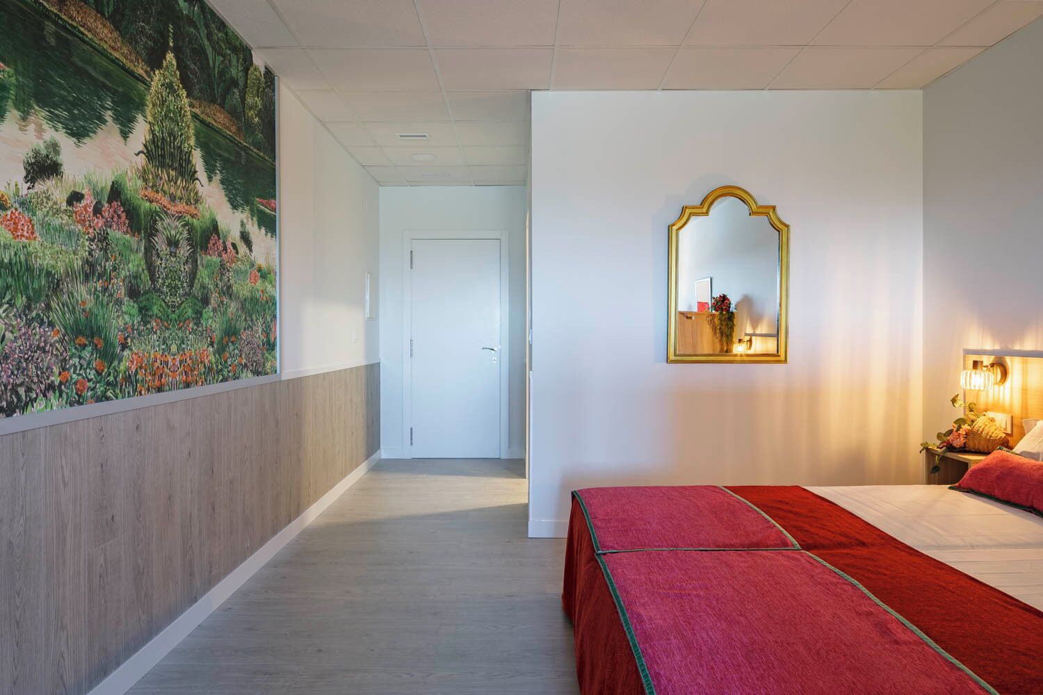 A hotel room with a bed, a mirror, a nightstand, and a wall with a floral painting.