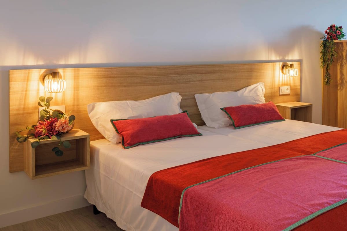 A neatly made bed with white linens and red accents, wooden headboard, and bedside tables with wall-mounted lamps.