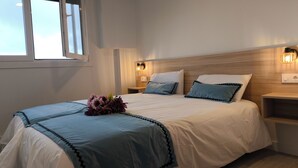 Una cama bien hecha con colcha azul y blanca, un arreglo floral decorativo y un cabecero de madera. Se observa una mesita de noche con una lámpara y un aplique de pared.