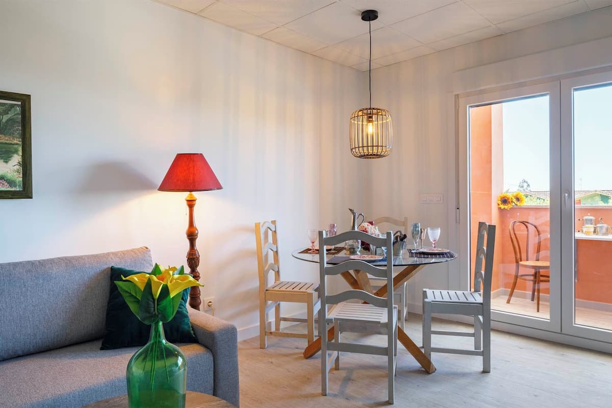 A living room with a sofa, a table with chairs, a lamp, and a view of the outside.